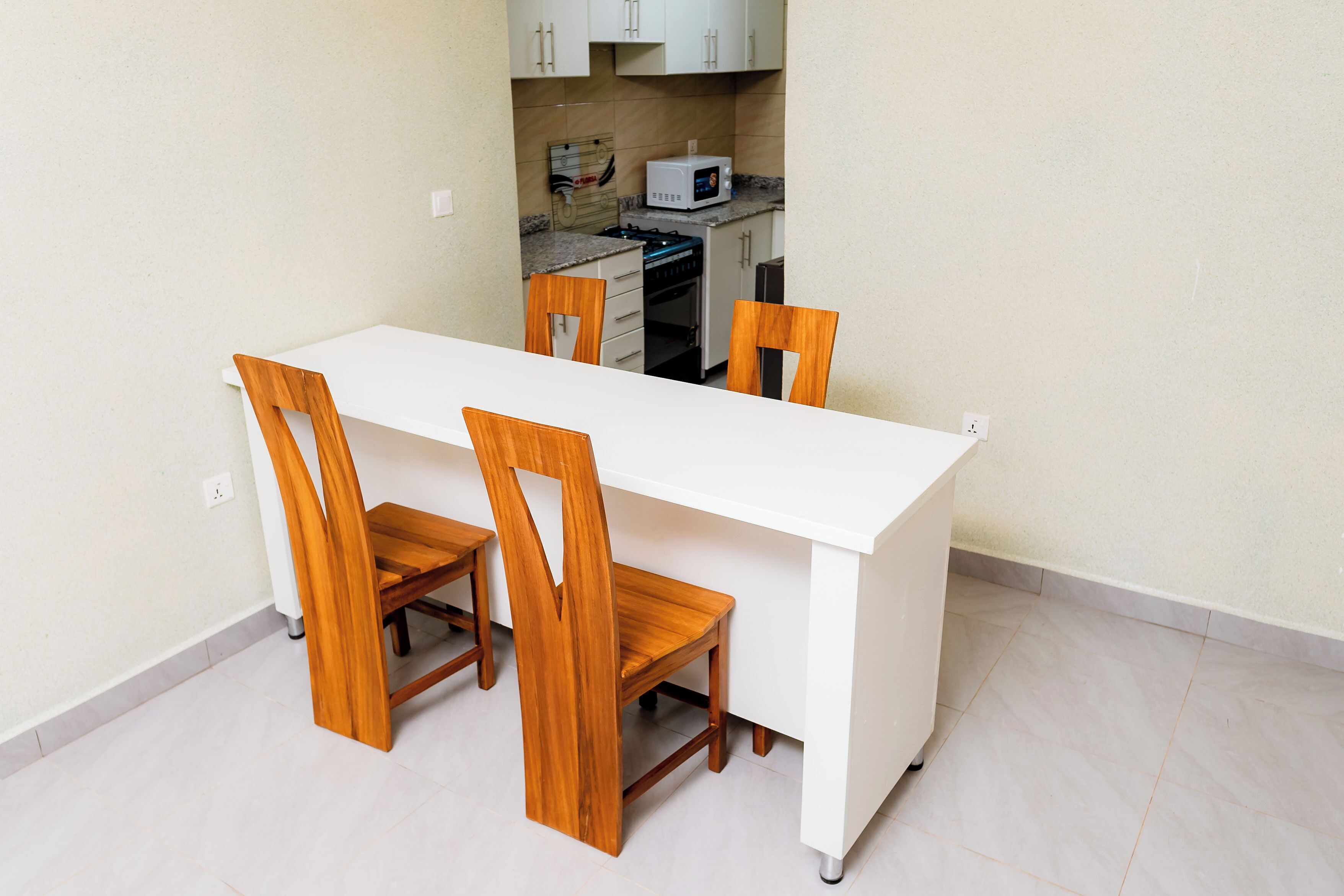A dining area with a white table and wooden chairs, a kitchen in the background, and a tiled floor.