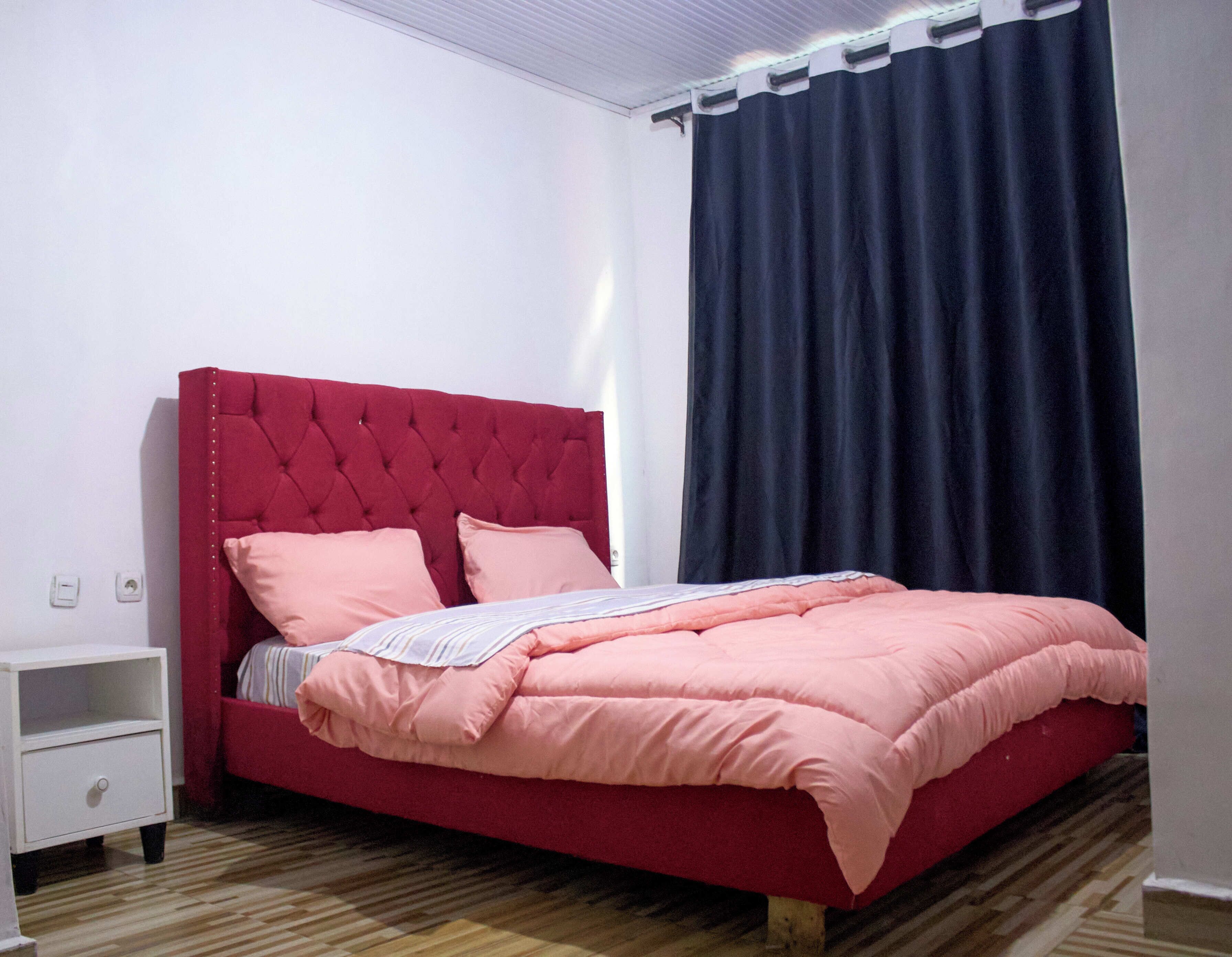 A neatly made bed with a red headboard and pink bedding, a white nightstand, and a dark curtain.