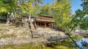 A cabin with a wooden deck and stairs leading to a balcony, surrounded by trees and a body of water. - Lakefront Fun in Sunrise Beach! Pet-friendly Home! (Sunrise Beach)