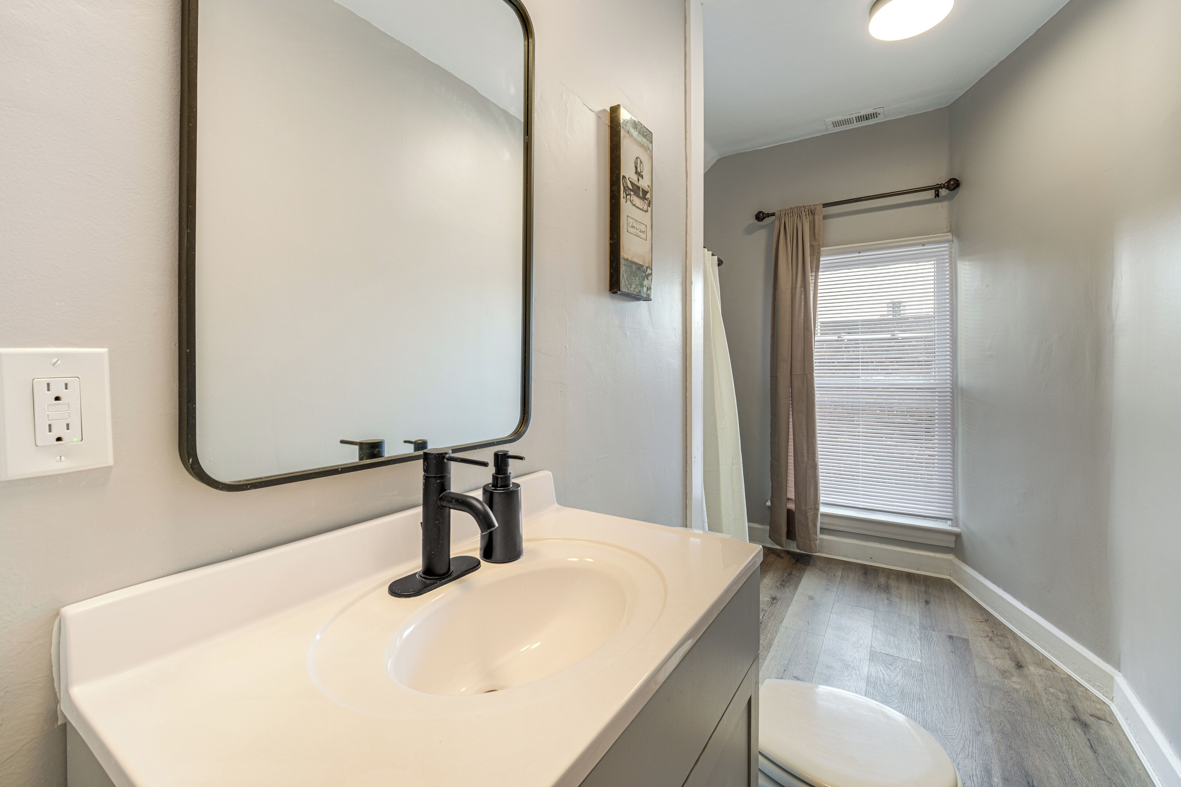 A bathroom with a white sink, black faucet, and a mirror. There is a window with blinds and a shower curtain.