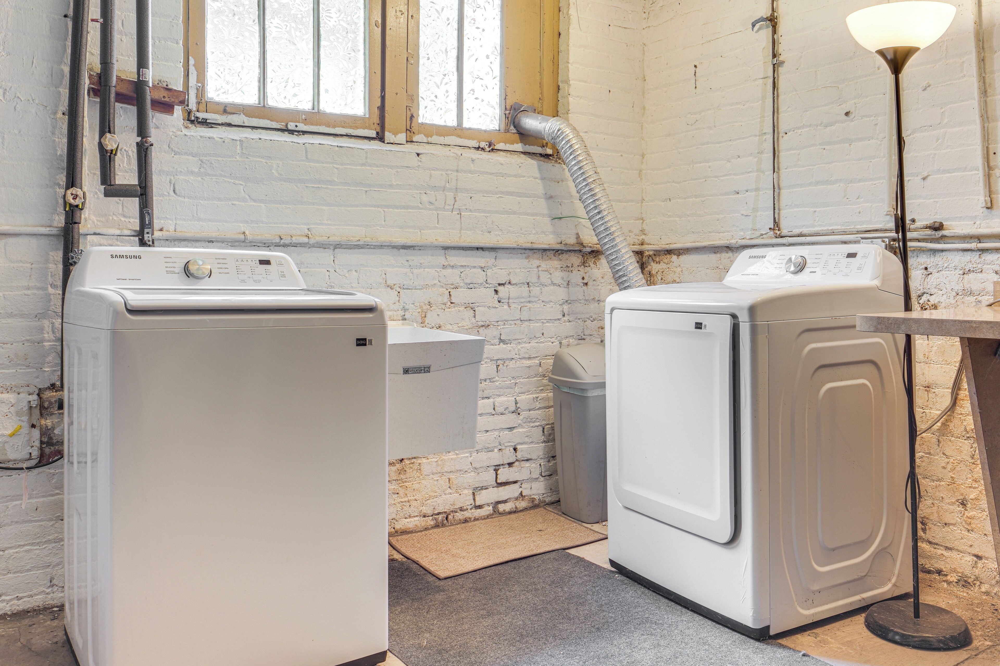 A laundry room with two white washing machines, a trash bin, and a floor lamp.