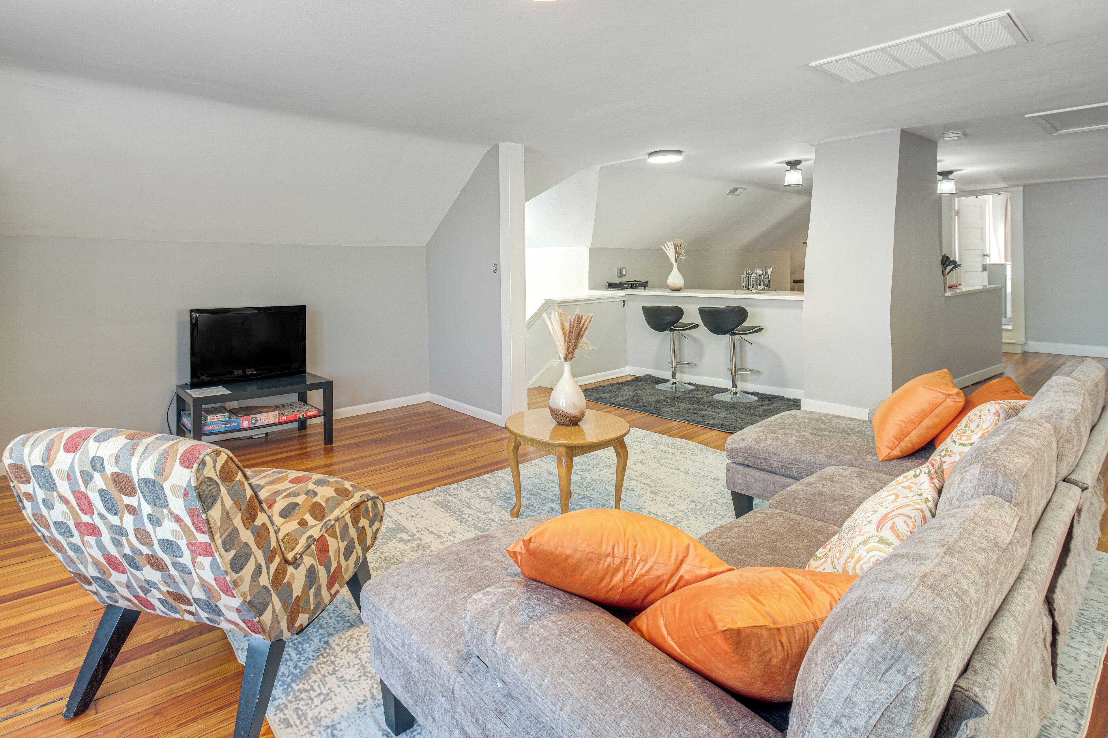 A modern living room with a grey sofa, a patterned armchair, a television, and a small dining area with stools.