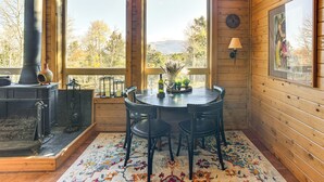 A cozy cabin interior with a fireplace, wooden walls, a dining table with chairs, and large windows offering a view of trees and mountains. - Fire Pit & Peak Views: Gold Camp Area Cabin (Cripple Creek)