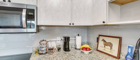 Une cuisine avec des meubles blancs, un four à micro-ondes, une machine à café, un saladier rempli de fruits et un tableau encadré.