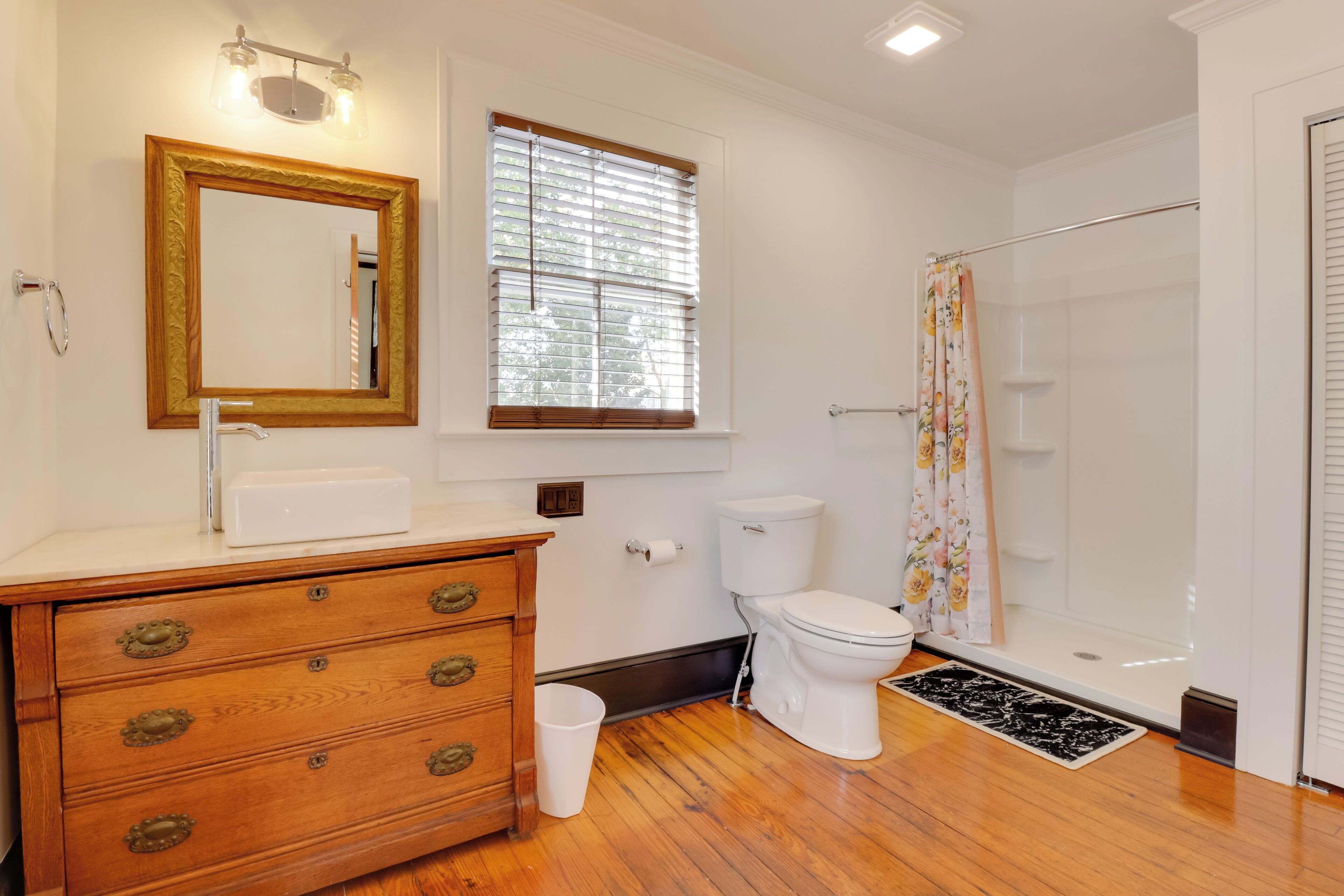 A bathroom with a wooden vanity, a toilet, a shower, and a window with blinds.