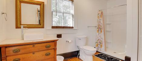 A bathroom with a wooden vanity, a toilet, a shower, and a window with blinds.