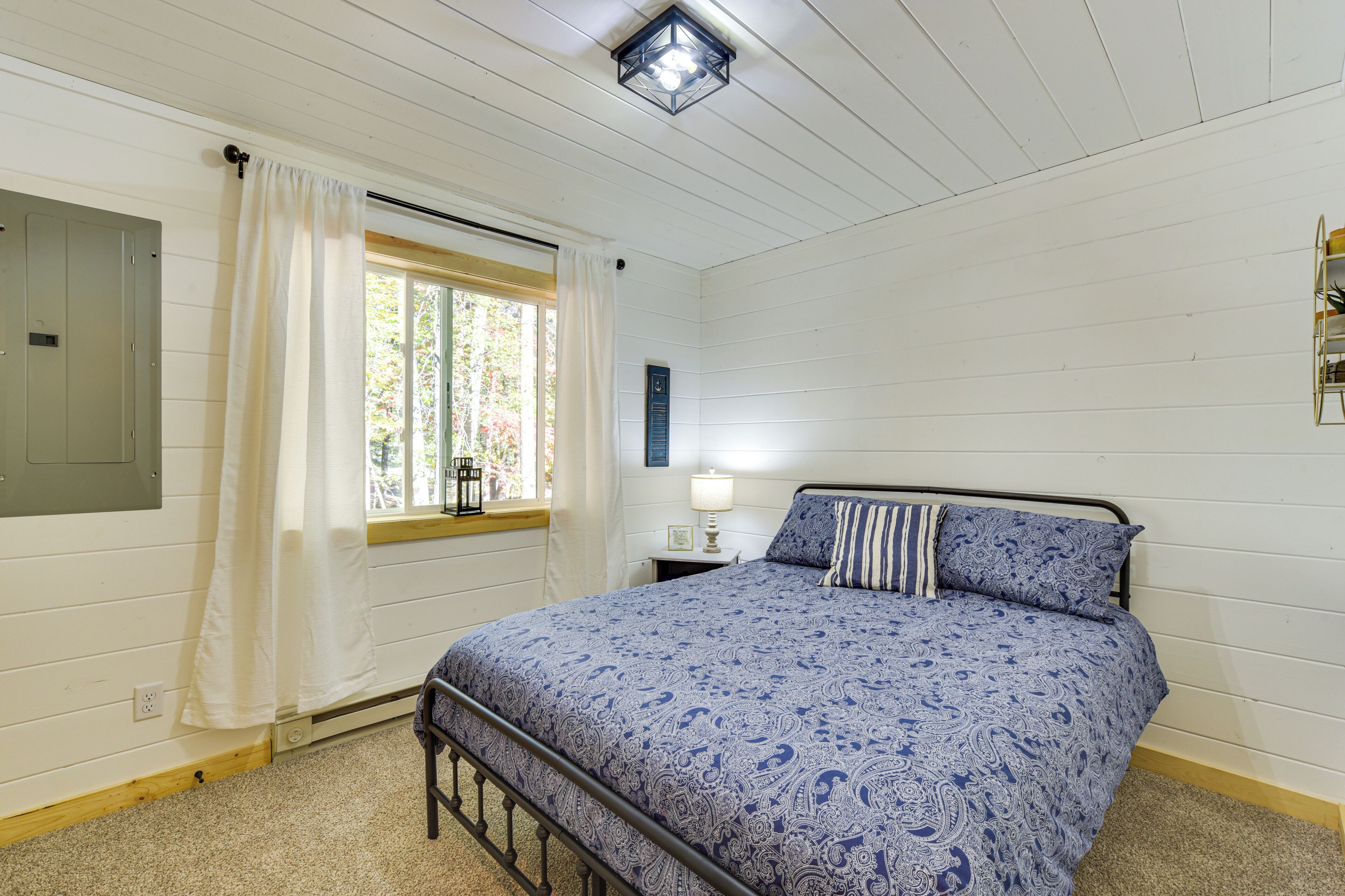 Ein Schlafzimmer mit einem Bett, einem Fenster mit Vorhängen, einem Nachttisch mit Lampe und einem an der Wand montierten Regal.