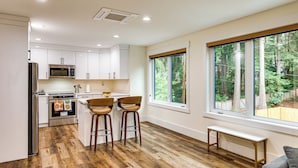 Una cocina moderna con armarios blancos, electrodomésticos de acero inoxidable y suelo de madera. Hay dos taburetes de bar y un banco cerca de un gran ventanal con vistas a un jardín.