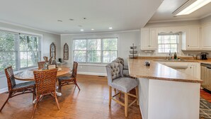 A spacious kitchen with white cabinetry, a large island, and a dining area with wicker chairs. - Walk to Dtwn Hahira: Inviting Home w/ Pool & Yard! (Hahira)
