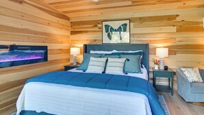 A bedroom with a wooden ceiling, a bed with blue bedding, two bedside tables with lamps, and a framed butterfly artwork above the bed. - Private Hot Tub, Fire Pit! Pickwick Lake Escape (Cherokee)