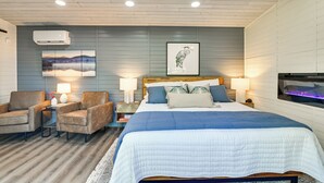 A hotel room with a bed, two armchairs, a side table with a lamp, a wall-mounted air conditioner, and a framed picture of a bird.