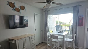 A room with a ceiling fan, a flat-screen TV, a dining table with chairs, a wooden cabinet, and a sliding glass door leading to an outdoor area. - Turtle Cove - Cozy retreat steps from the ocean, (Kure Beach)