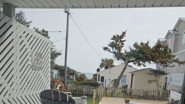 A deck with two chairs, a table, and a view of a fenced yard with a house and trees. - Turtle Cove - Cozy retreat steps from the ocean, (Kure Beach)