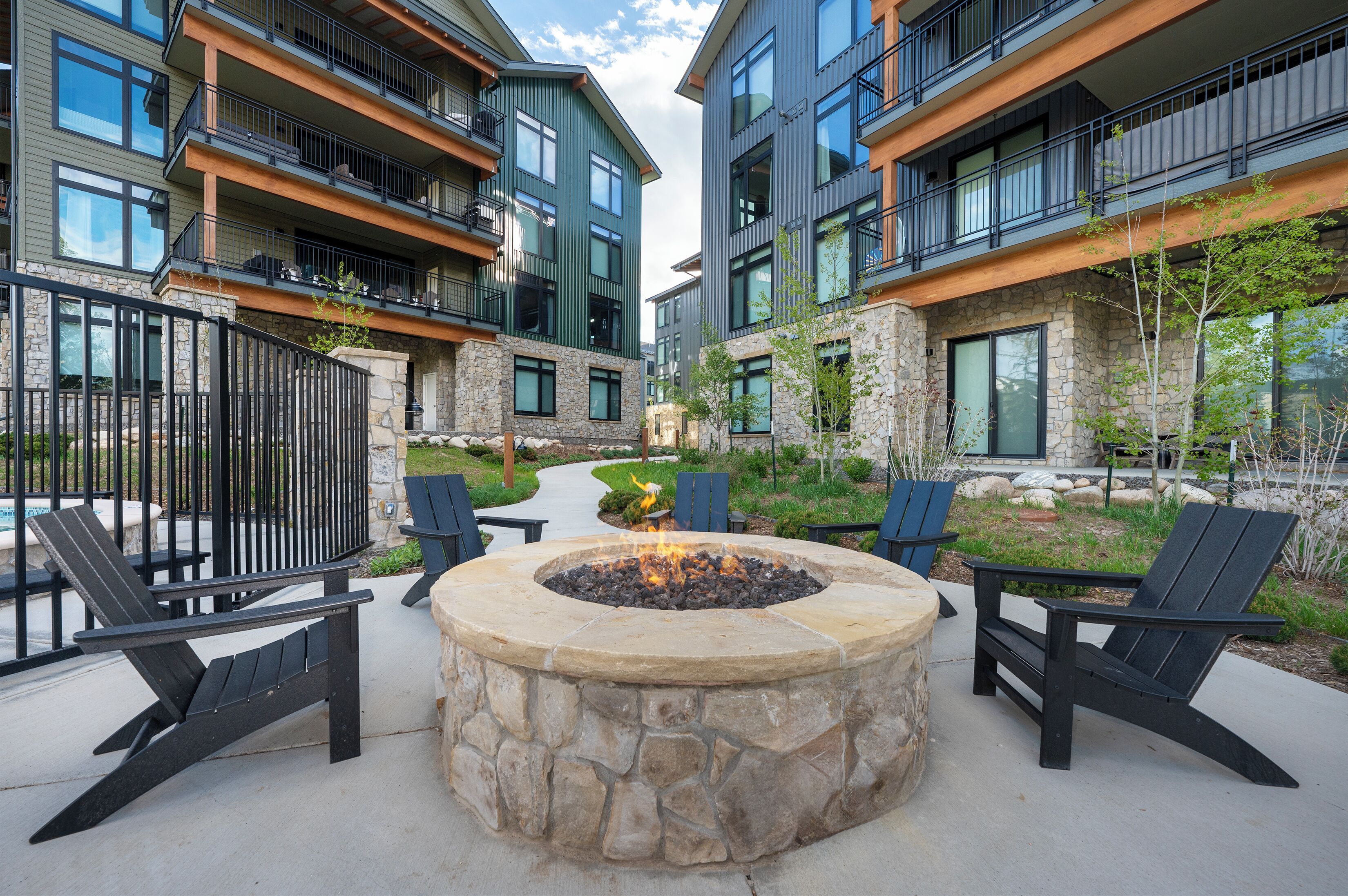 A courtyard with a stone fire pit surrounded by Adirondack chairs, modern multi-story buildings, and a stone pathway.