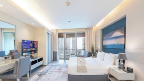 A modern hotel room with a large bed, a desk with a TV, a balcony with a city view, and a large painting on the wall. - PVH - Emaar Residences - Studio (Dubai)