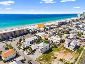 Vue aérienne d’une ville côtière avec un hôtel en bord de mer, comprenant un parking, des bâtiments et une vue dégagée sur la plage et l’océan.