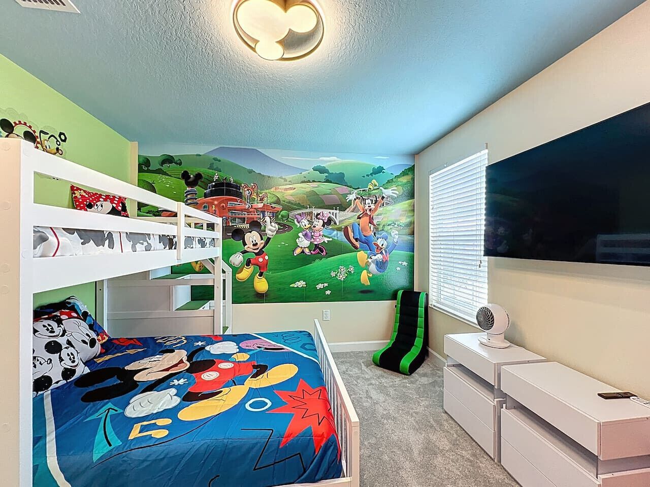 Une chambre d’enfant avec des lits superposés, une fresque murale, un téléviseur et un bureau avec des tiroirs.