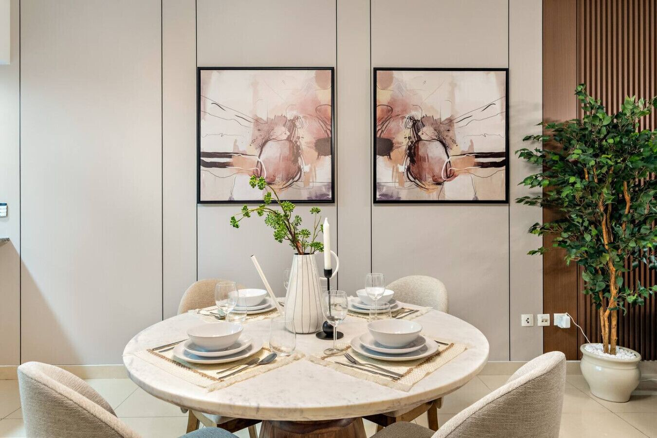 A dining area with a round table set for four, two framed abstract paintings on the wall, a potted plant, and a wooden paneling design.