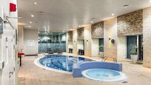 A modern indoor pool area with a large rectangular pool, a smaller circular hot tub, and a seating area with tables and chairs.