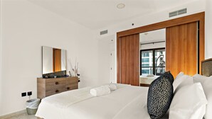 A modern bedroom with a large bed, wooden headboard, a vanity with a mirror, and a view of the city through a window.