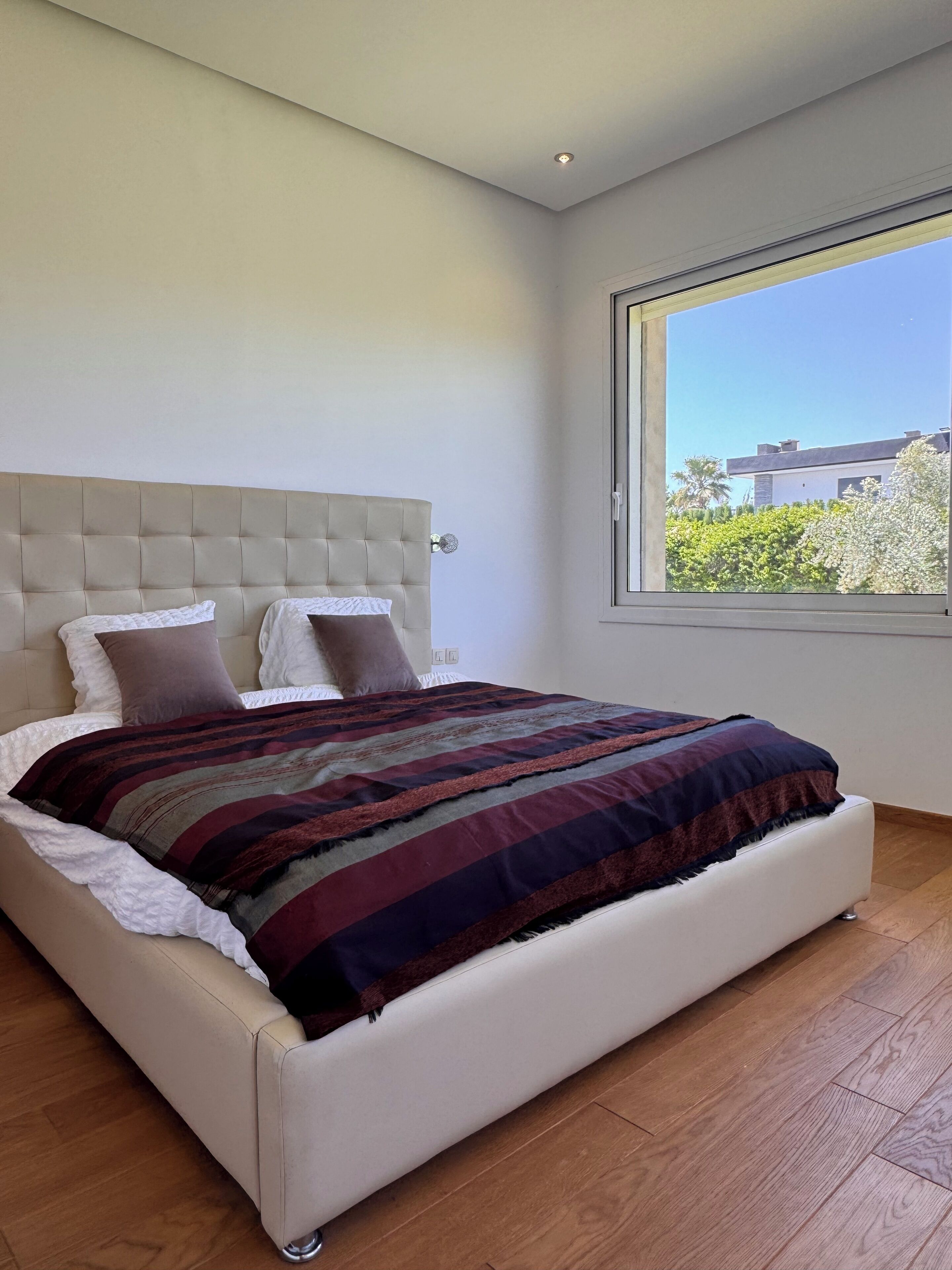 A modern bedroom with a large bed, a tufted headboard, and a view of a building through a window.