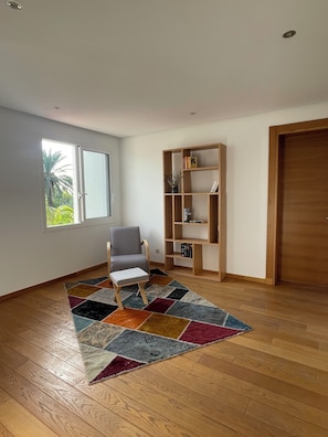 A room with a wooden floor, a grey armchair, a bookshelf, and a window with a view of greenery.