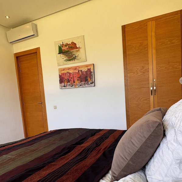 A bedroom with a bed, two wooden wardrobes, an air conditioner, and two paintings on the wall.