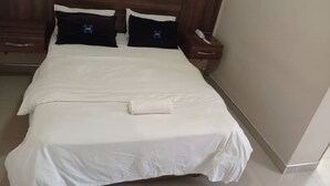 A neatly made bed with white bedding and two black pillows with a logo, a wooden headboard, a bedside table with a phone, and a drawer unit. - North Stay Inn (Livingstone)