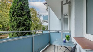 Ein Balkon mit Tisch und Stühlen, einem Blumentopf und Blick auf Bäume und Gebäude.