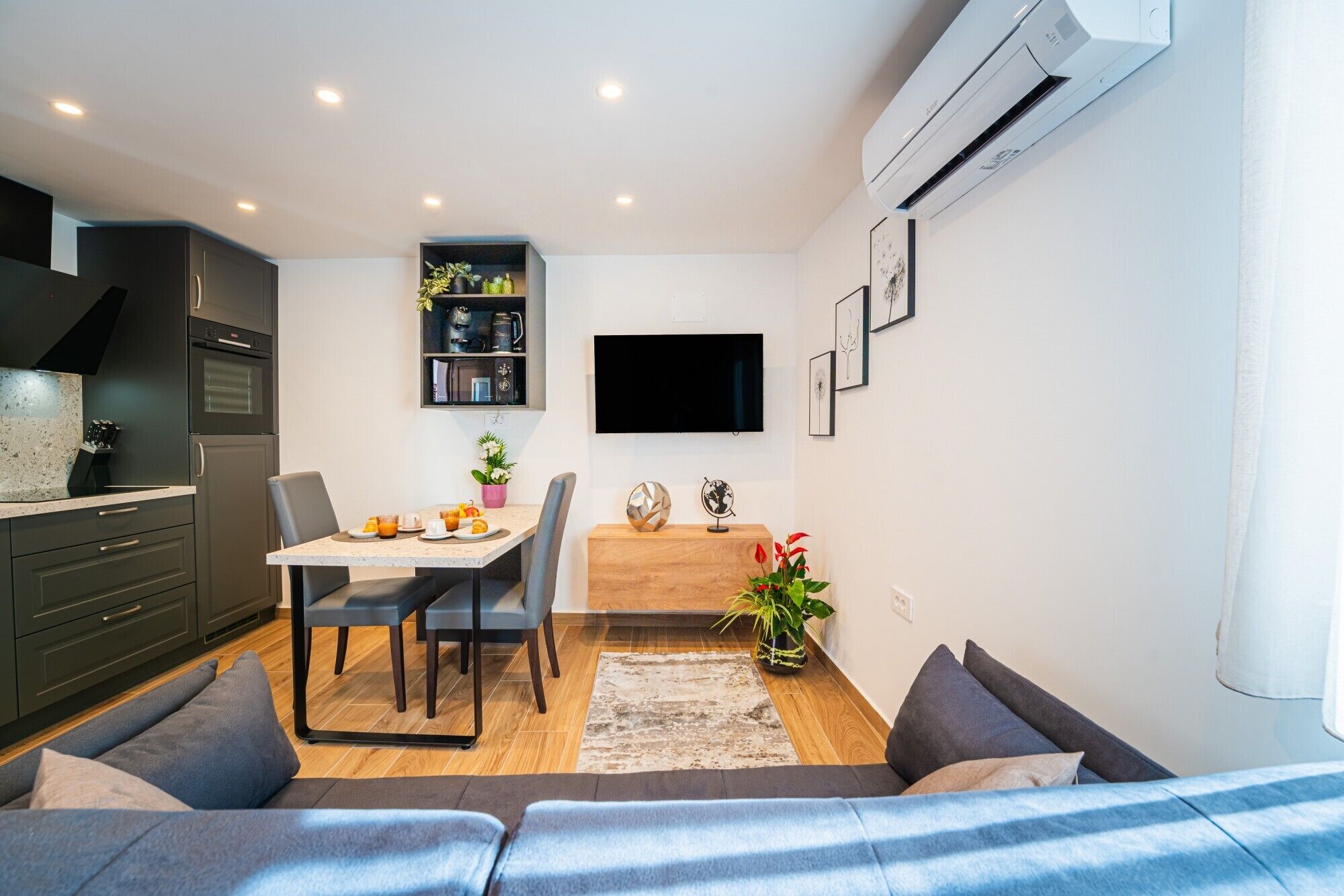 Eine moderne Küche mit dunklen Schränken, eine Essecke mit Tisch und Stühlen, ein an der Wand befestigter Flachbildfernseher und eine eingebaute Klimaanlage.