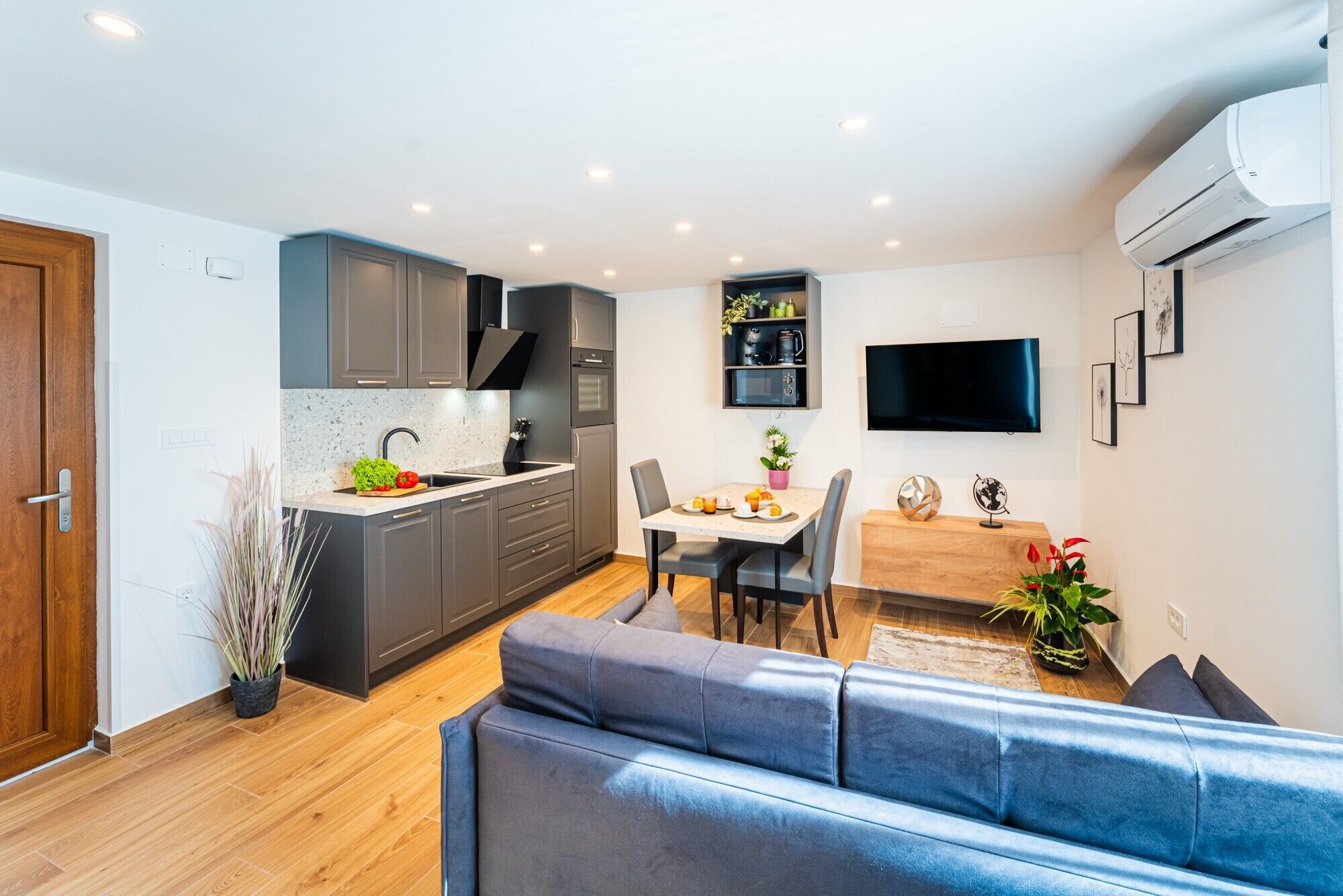 Een moderne keuken met grijze kastjes, een eethoek met tafel en stoelen, en een zithoek met een blauwe bank.