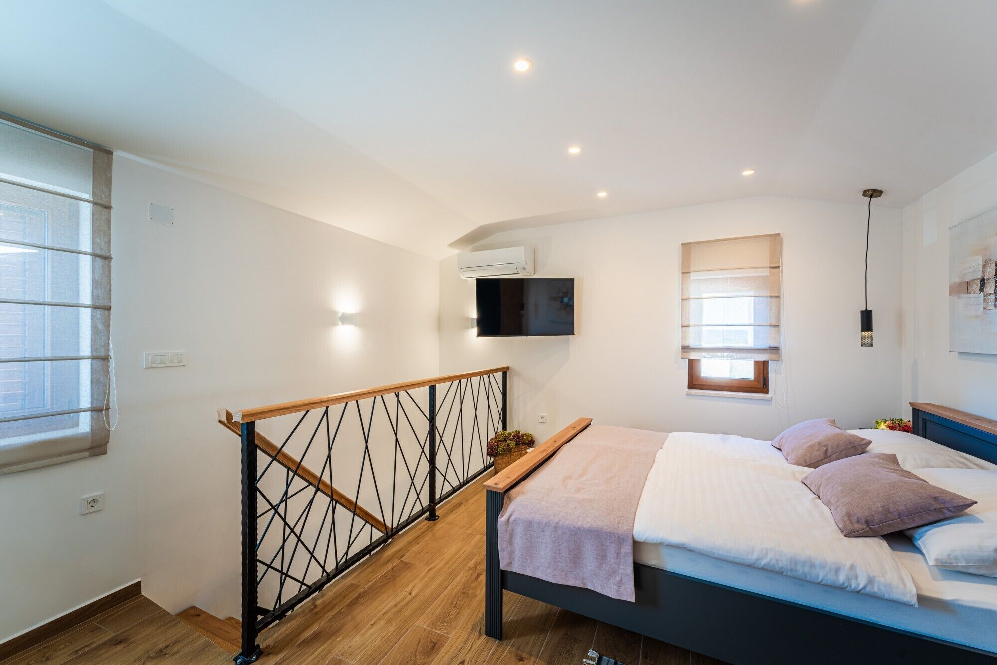 Une chambre à coucher comprenant un lit, un téléviseur fixé au mur, une fenêtre avec des stores et un escalier doté d’une rampe en métal.