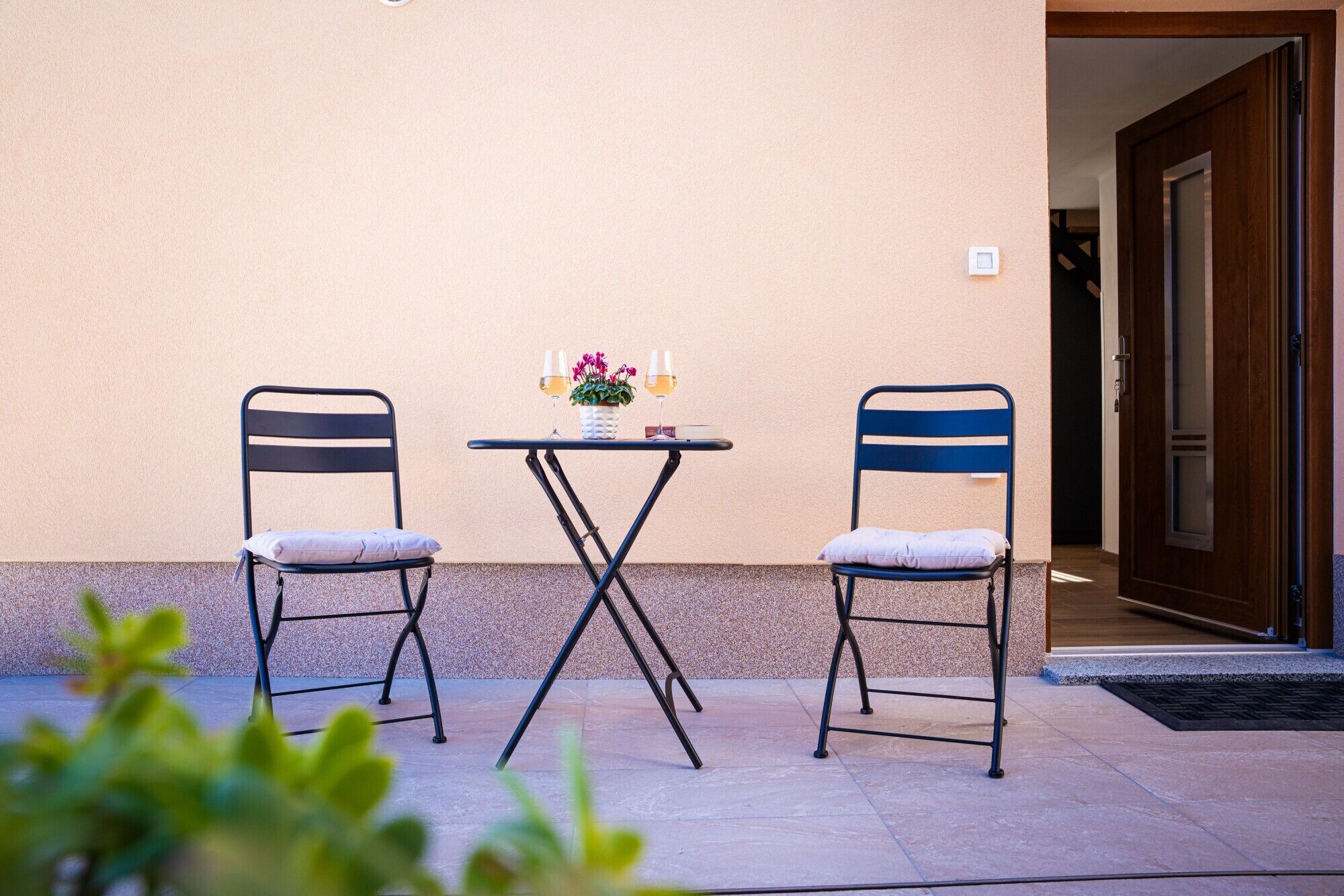 Two chairs and a small table with a potted plant and two glasses of wine against a beige wall.