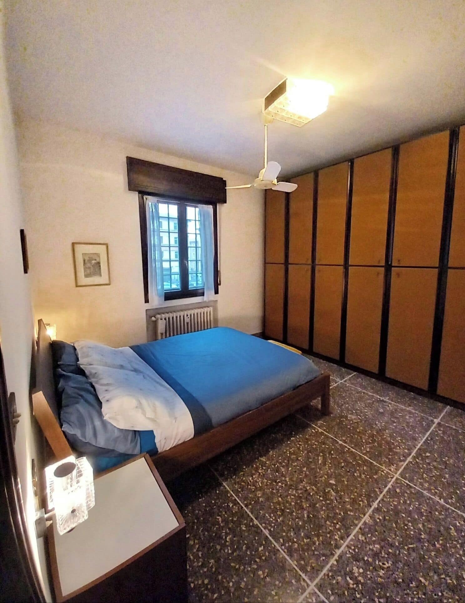 Una camera da letto con un letto, una finestra, un termosifone, una ventola a soffitto e un armadio.