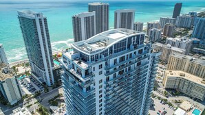 Un hotel di grandi dimensioni con una terrazza panoramica sul tetto, circondato da altri edifici alti e una spiaggia sullo sfondo.