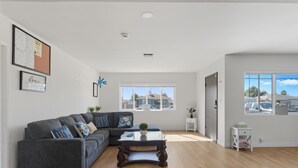 A spacious living room with a grey sectional sofa, a glass coffee table, and a view of the neighborhood through large windows. - Relaxing Paradise Hills Retreat 4 Bedroom Home (San Diego)