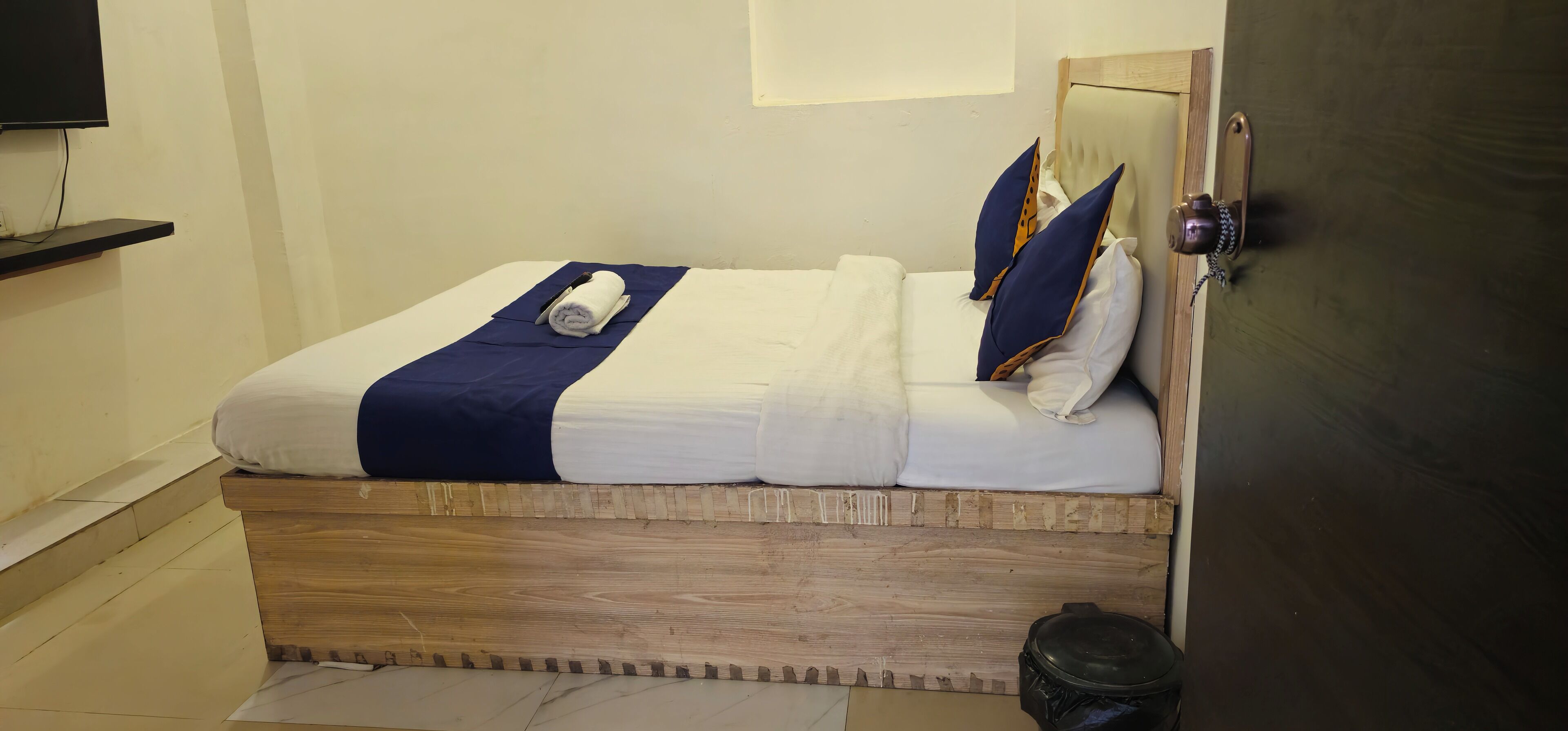 A neatly made bed with a blue blanket and pillows, a wooden headboard, and a small trash bin beside the bed.