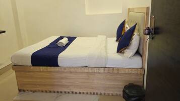 A neatly made bed with a blue blanket and pillows, a wooden headboard, and a small trash bin beside the bed.