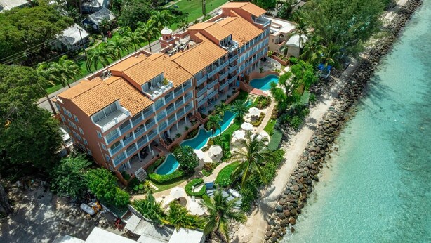 A multi-story hotel with a swimming pool, surrounded by palm trees and a beach. - Villas On The Beach 305 2 Bedroom Home (Holetown)