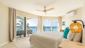 A bedroom with a bed, a nightstand, a ceiling fan, and a view of the ocean. - Villas On The Beach 305 2 Bedroom Home (Holetown)