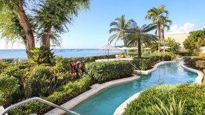 A pool surrounded by lush greenery, palm trees, and a view of the ocean with boats. - Villas On The Beach 305 2 Bedroom Home (Holetown)