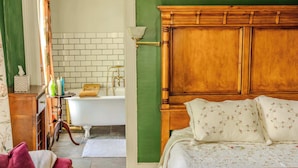 Un dormitorio con cama de madera, pared verde, baño con azulejos blancos y una silla roja.