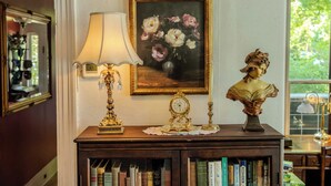 Una habitación con un aparador de madera, una lámpara, un cuadro de flores, un busto y una estantería llena de libros.