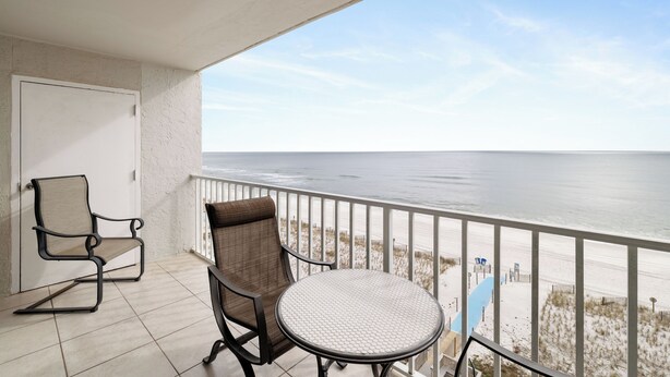 A balcony with two chairs and a table, overlooking a beach with a fence and beach chairs.