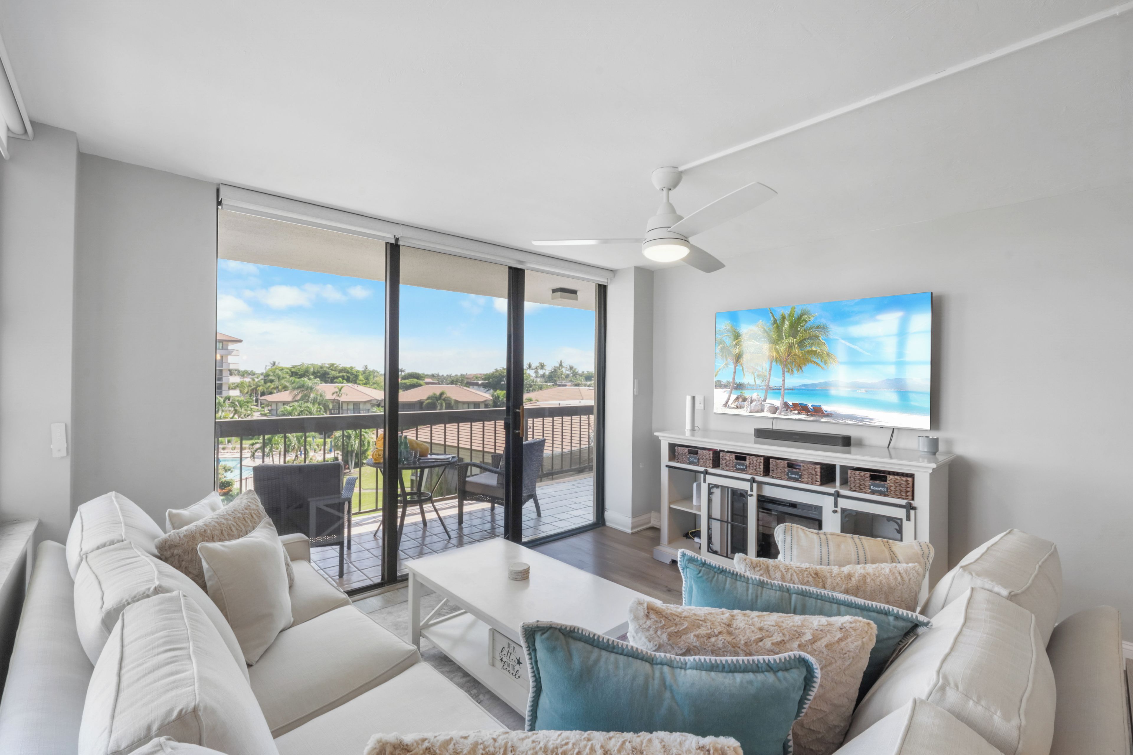 A modern living room with a white sofa, a coffee table, and a TV on a white stand. There is a balcony with a view of the city.