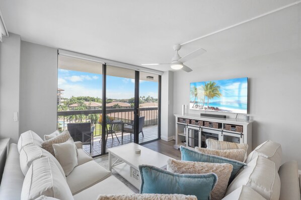 A modern living room with a white sofa, a coffee table, and a TV on a white stand. There is a balcony with a view of the city.