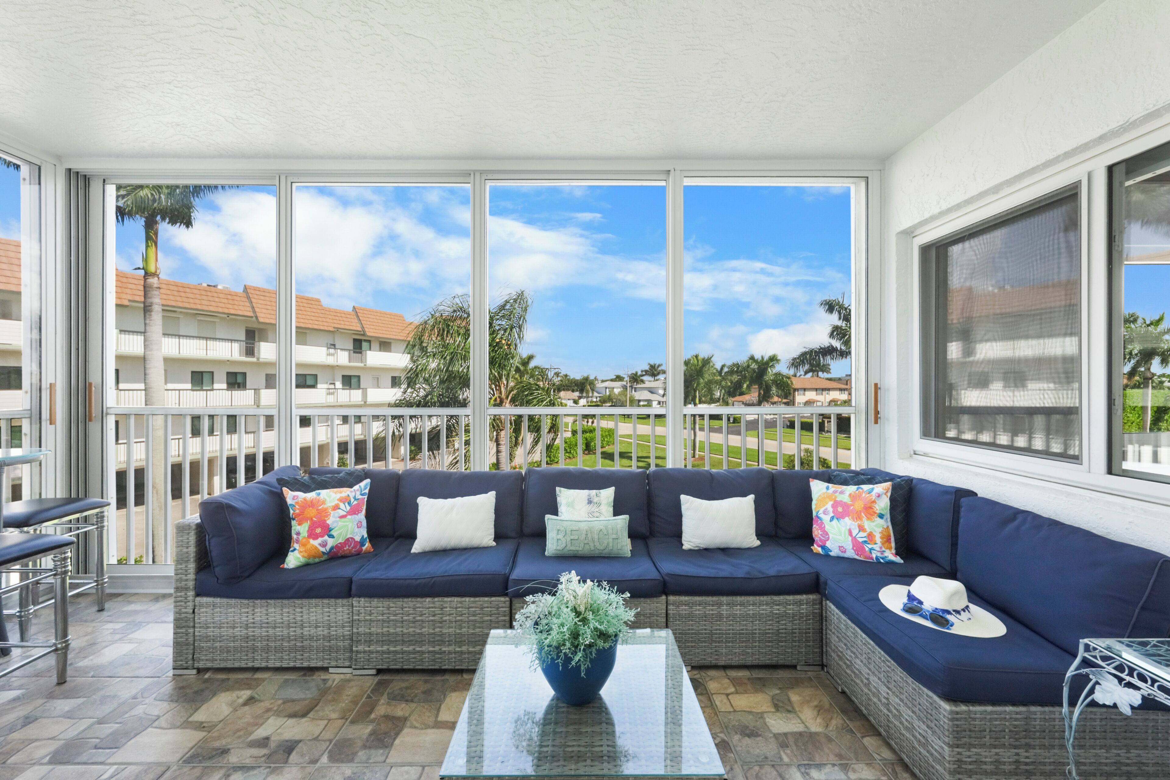 A balcony with a wicker sofa, a glass coffee table, and a view of buildings and palm trees.