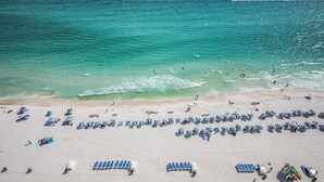 An aerial view of a beach with rows of sun loungers and umbrellas, a sandy shoreline, and the ocean.