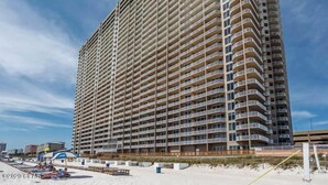 A high-rise hotel with multiple balconies, located near a beach with sun loungers and umbrellas.