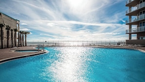 A swimming pool with a clear view of the ocean, surrounded by palm trees and a building with balconies.